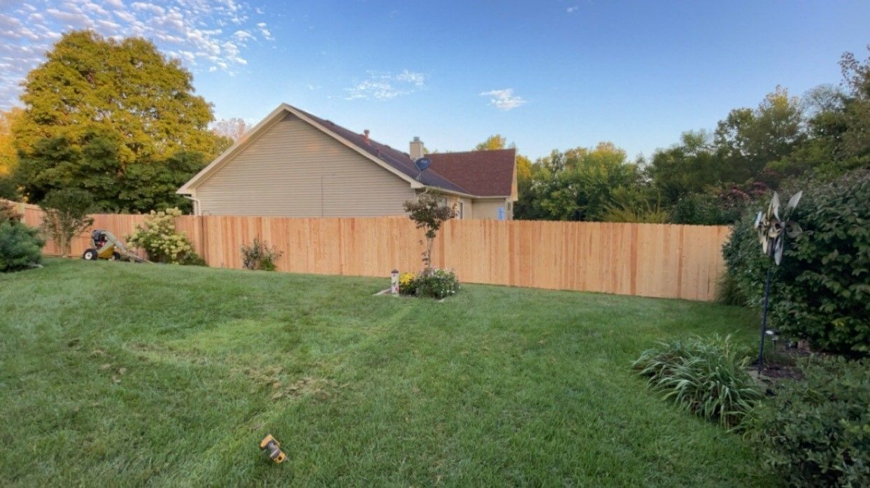 Backyard with a small beige house, wooden fence, and green lawn under a clear blue sky