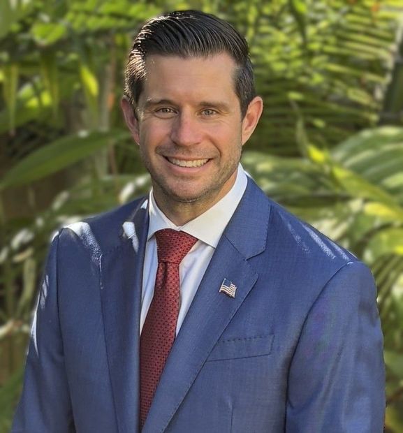 Smiling man in a blue suit and red tie standing outdoors with green foliage behind him