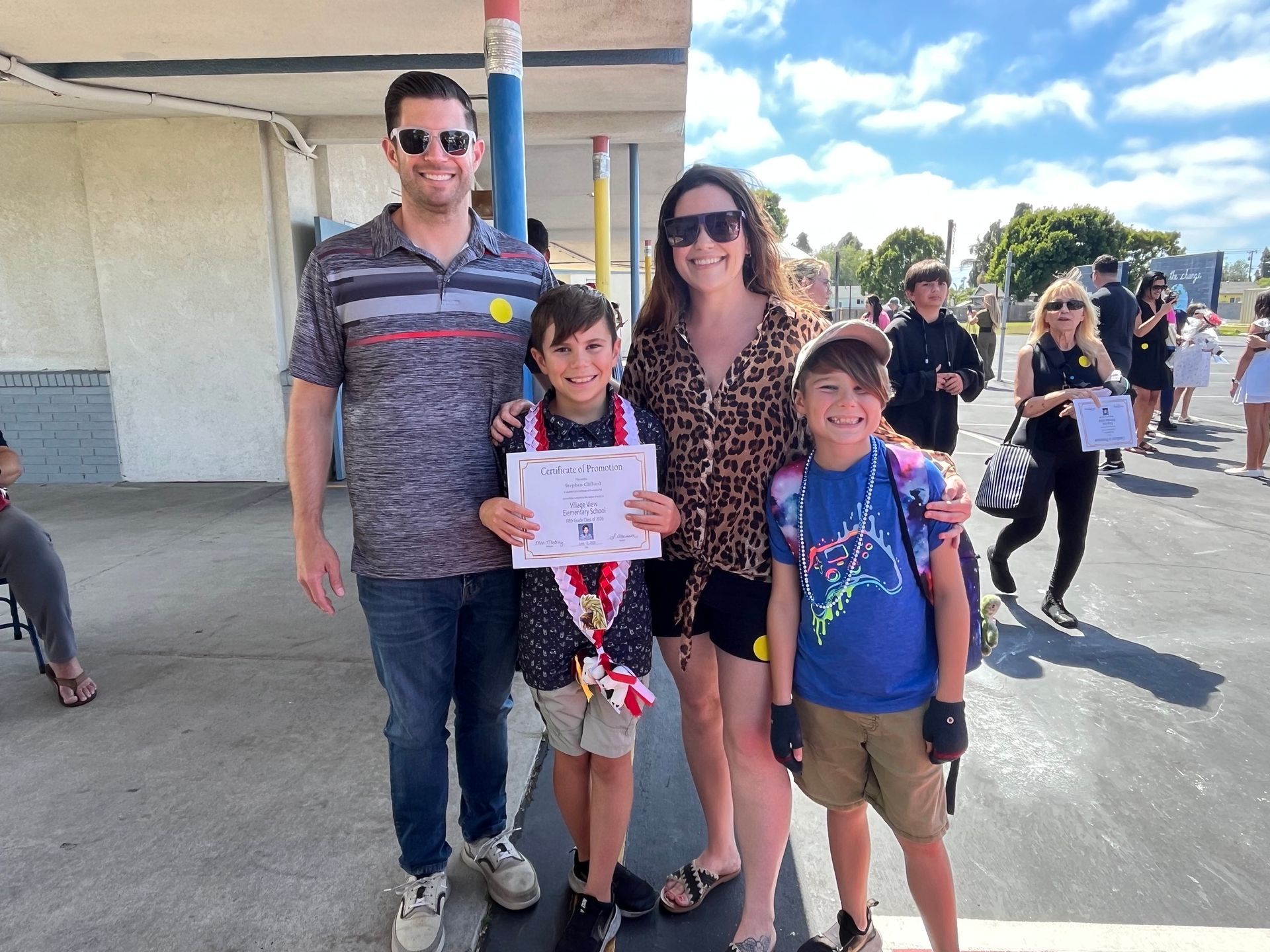 Family of four posing outdoors, with two children holding a certificate at a sunny event venue.