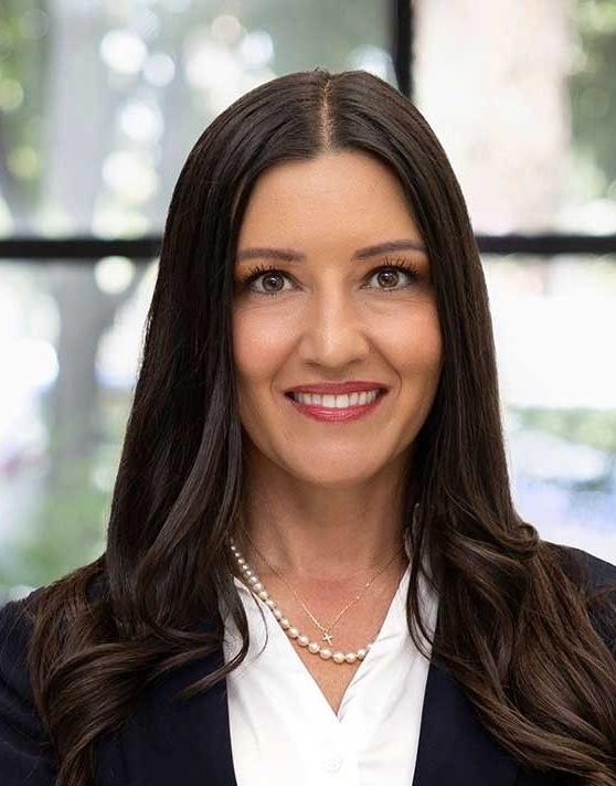 Sonja Shaw Portrait of a smiling professional in a white blouse and dark blazer against a bright windowed background