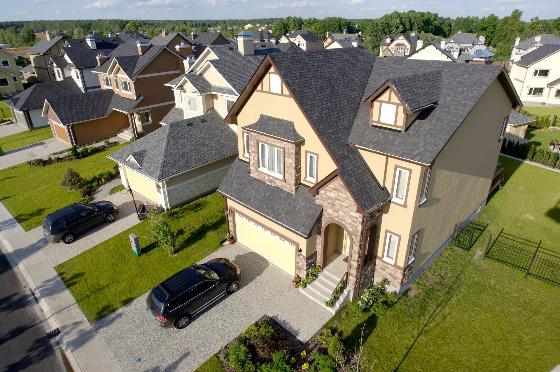 Suburban houses with green lawns and dark cars parked in driveways, sunny day.