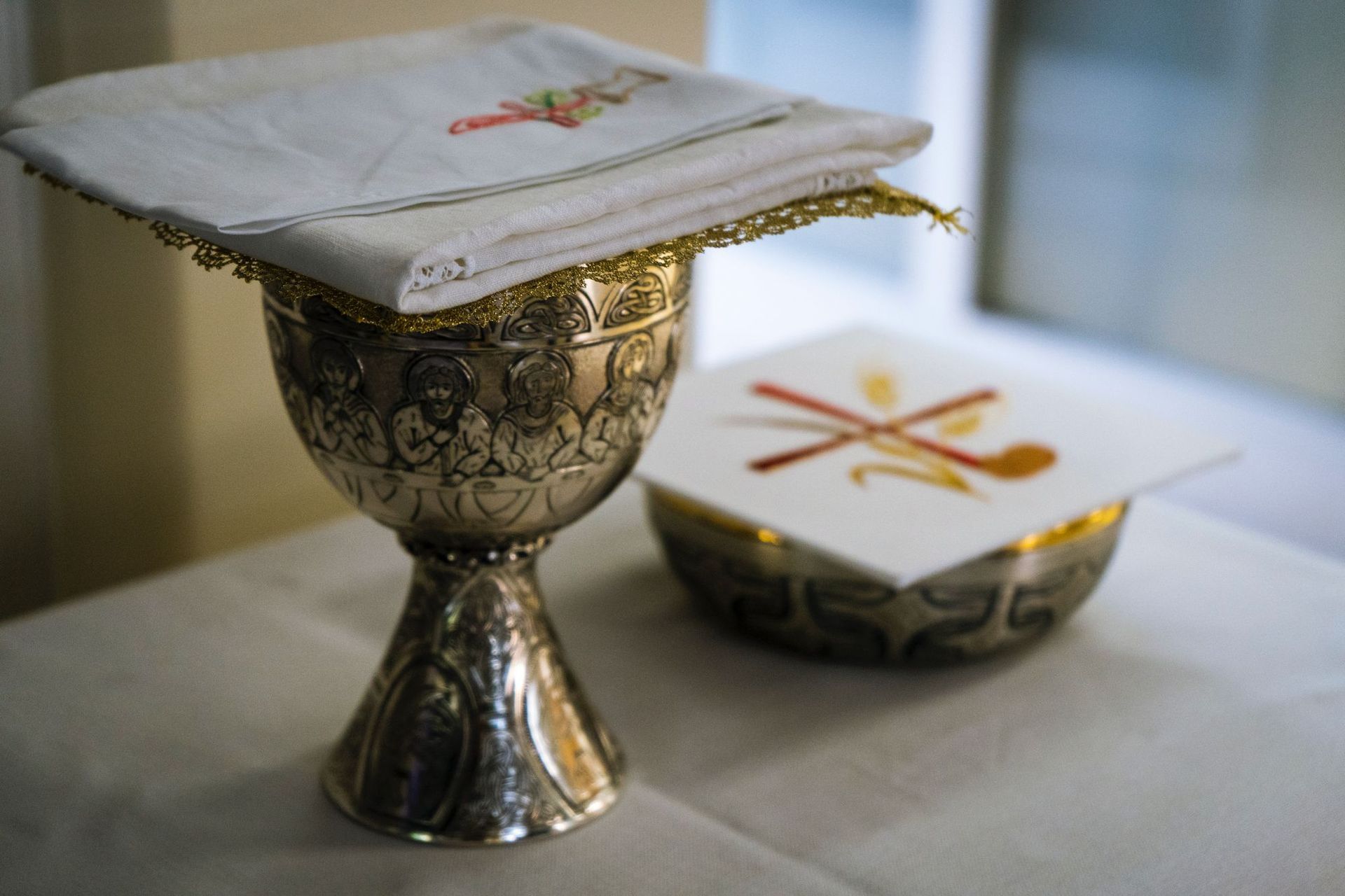 Silver chalice and covered dish on a table, with embroidered cloth draped over the chalice top.