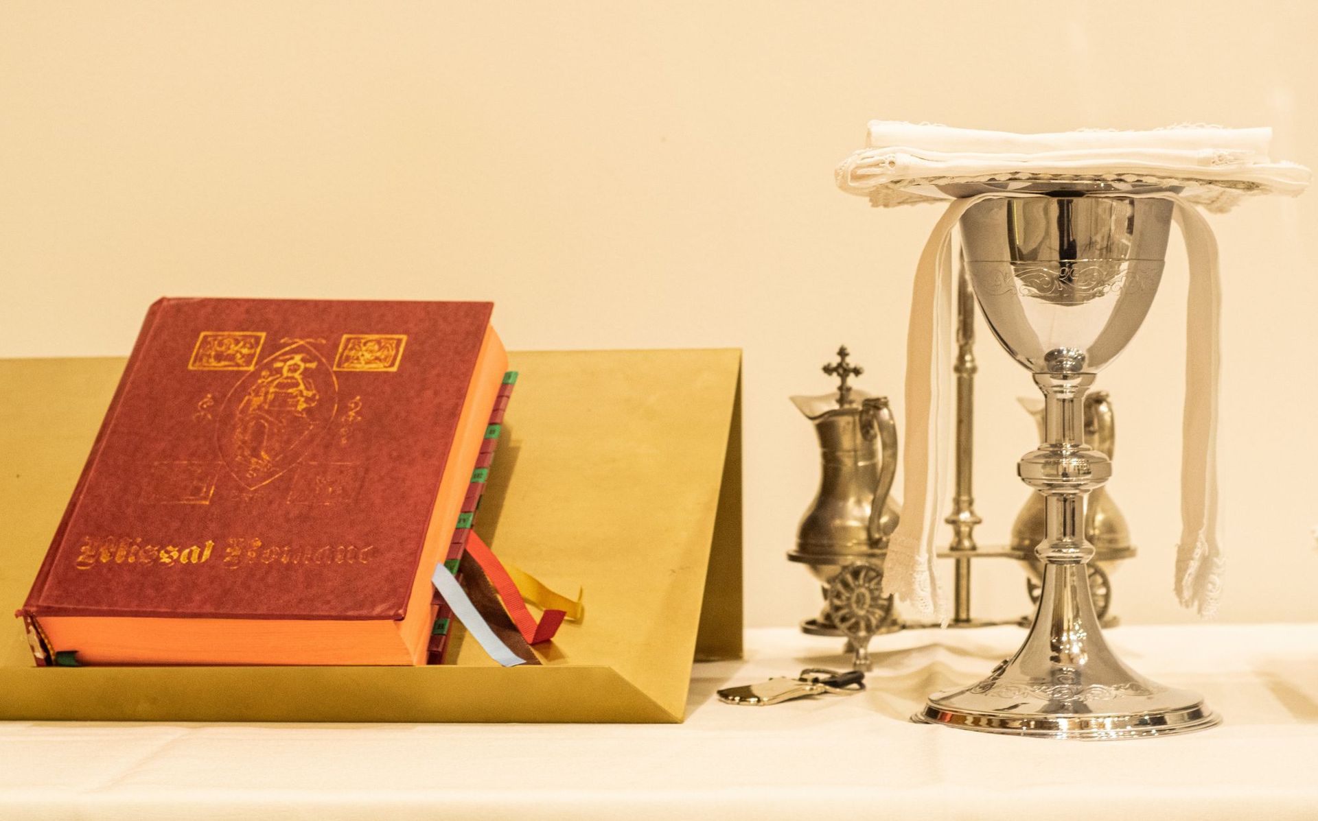 Red prayer book beside a silver chalice and candle on a white altar cloth