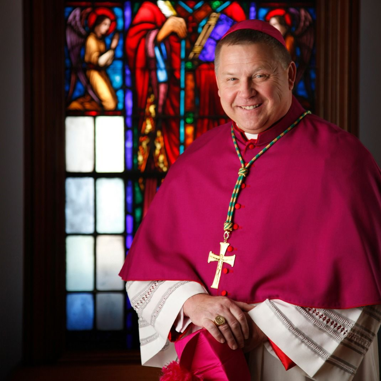 Archbishop Richard F. Stika in magenta robes smiles, holding a hat.  Window with stained glass is in the background.