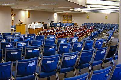 Rows of blue and red chairs face a podium, altar, and crucifix in a church meeting room.