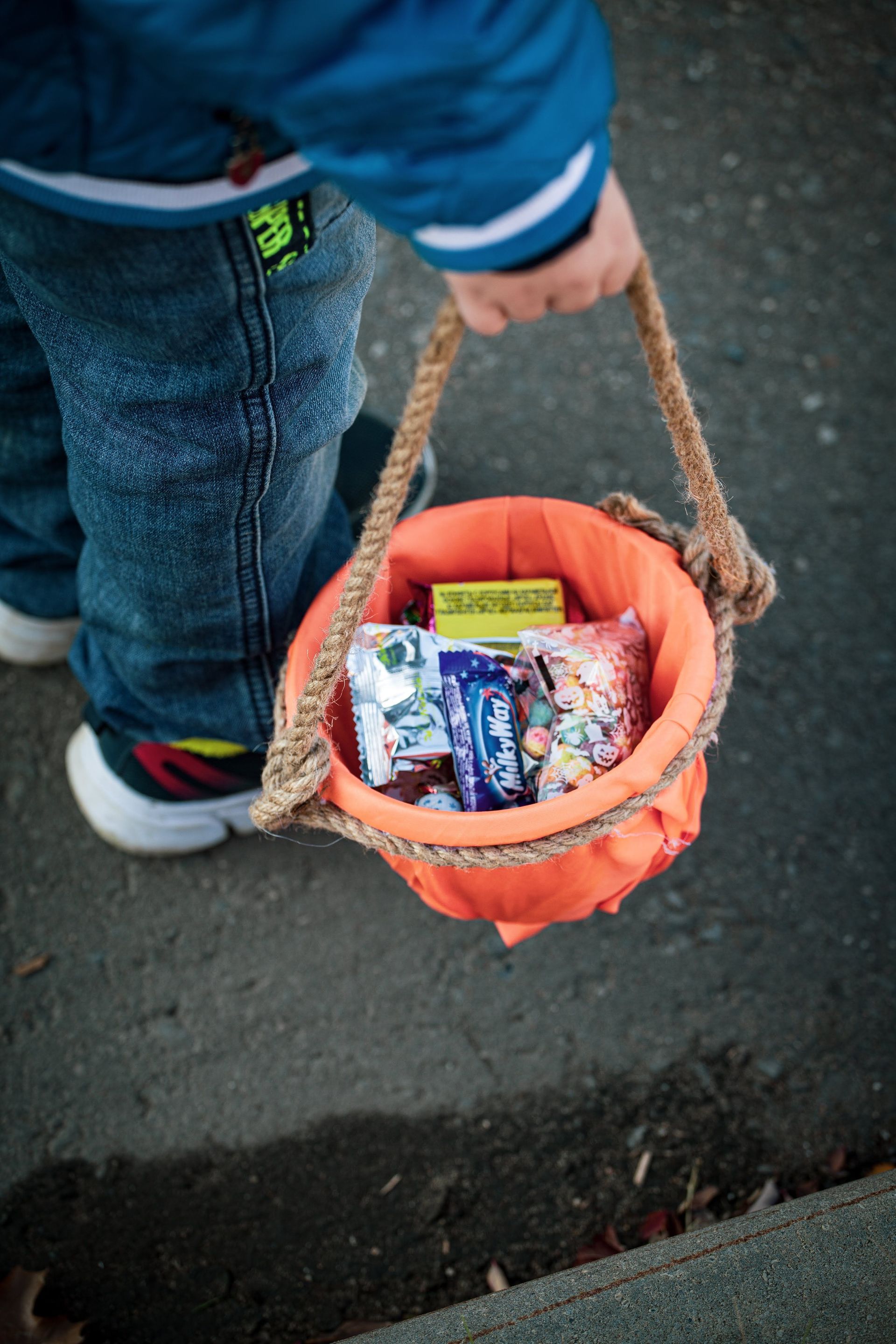 Child holding a small orange basket of candy on a sidewalk, with jeans and a blue jacket visible