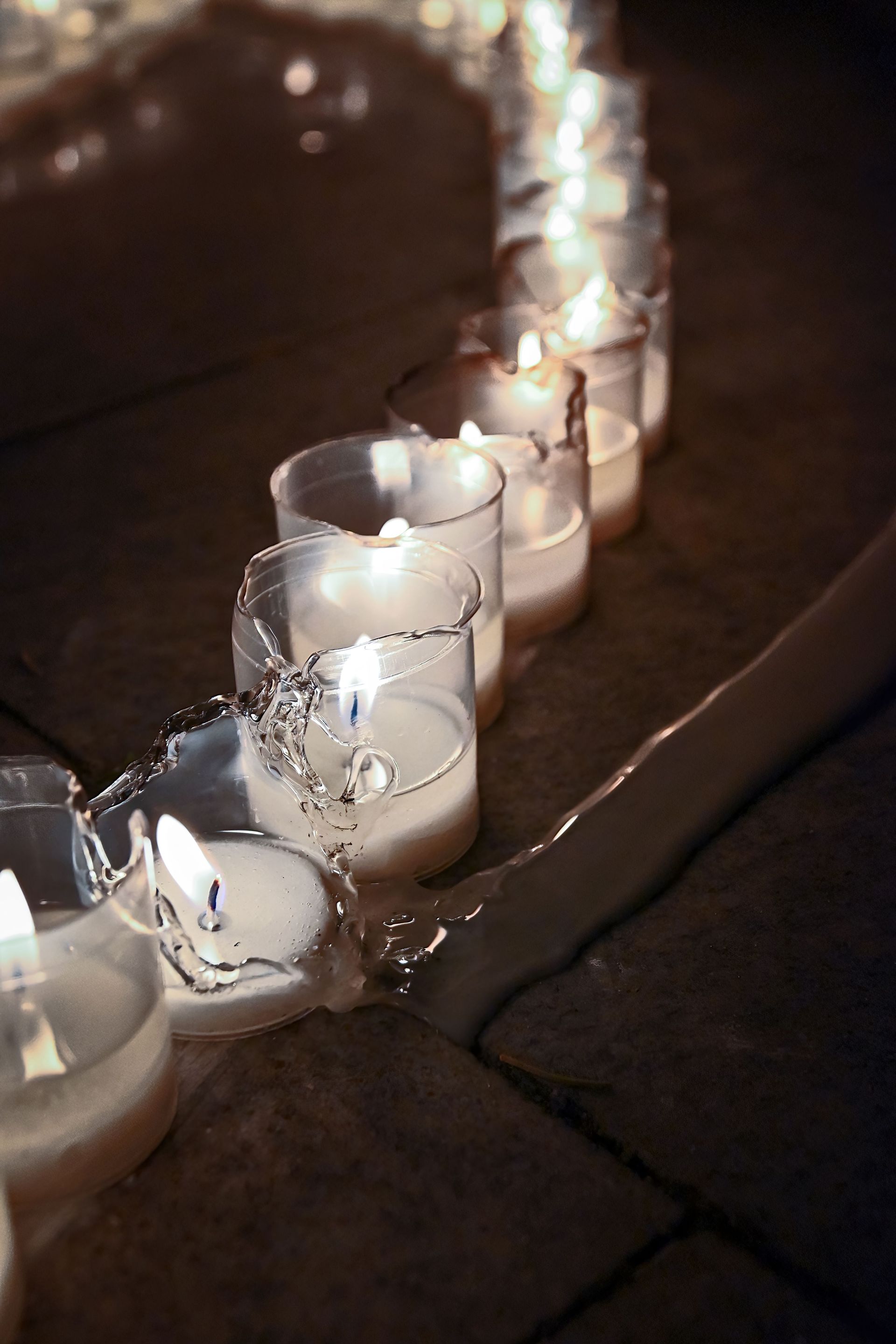 Small white candles glowing in glass holders along a wet sidewalk at night