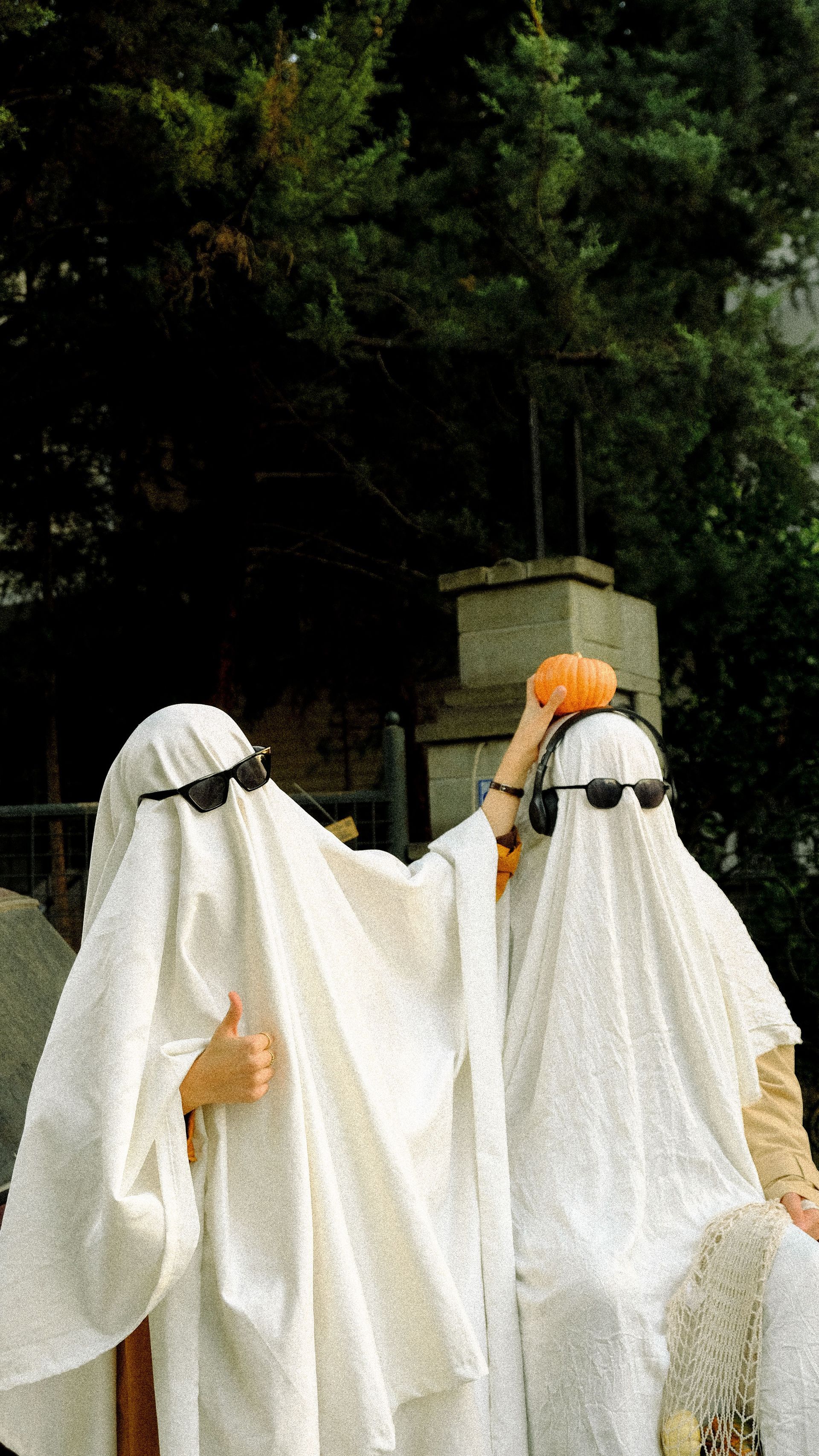 Two children in white ghost costumes standing outdoors near a tree and stone structure