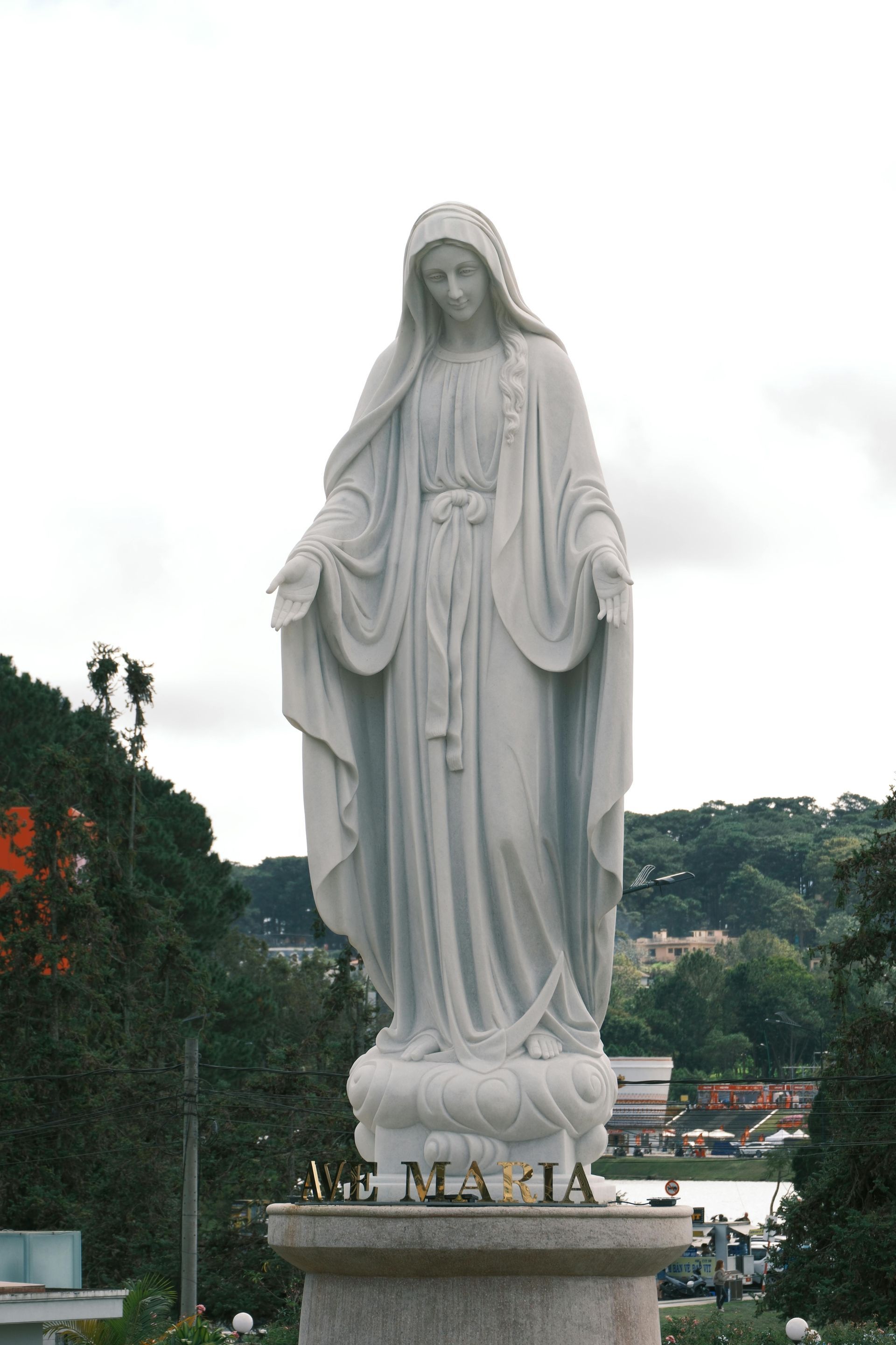 White statue of a robed figure with open arms on a pedestal against a cloudy hillside background