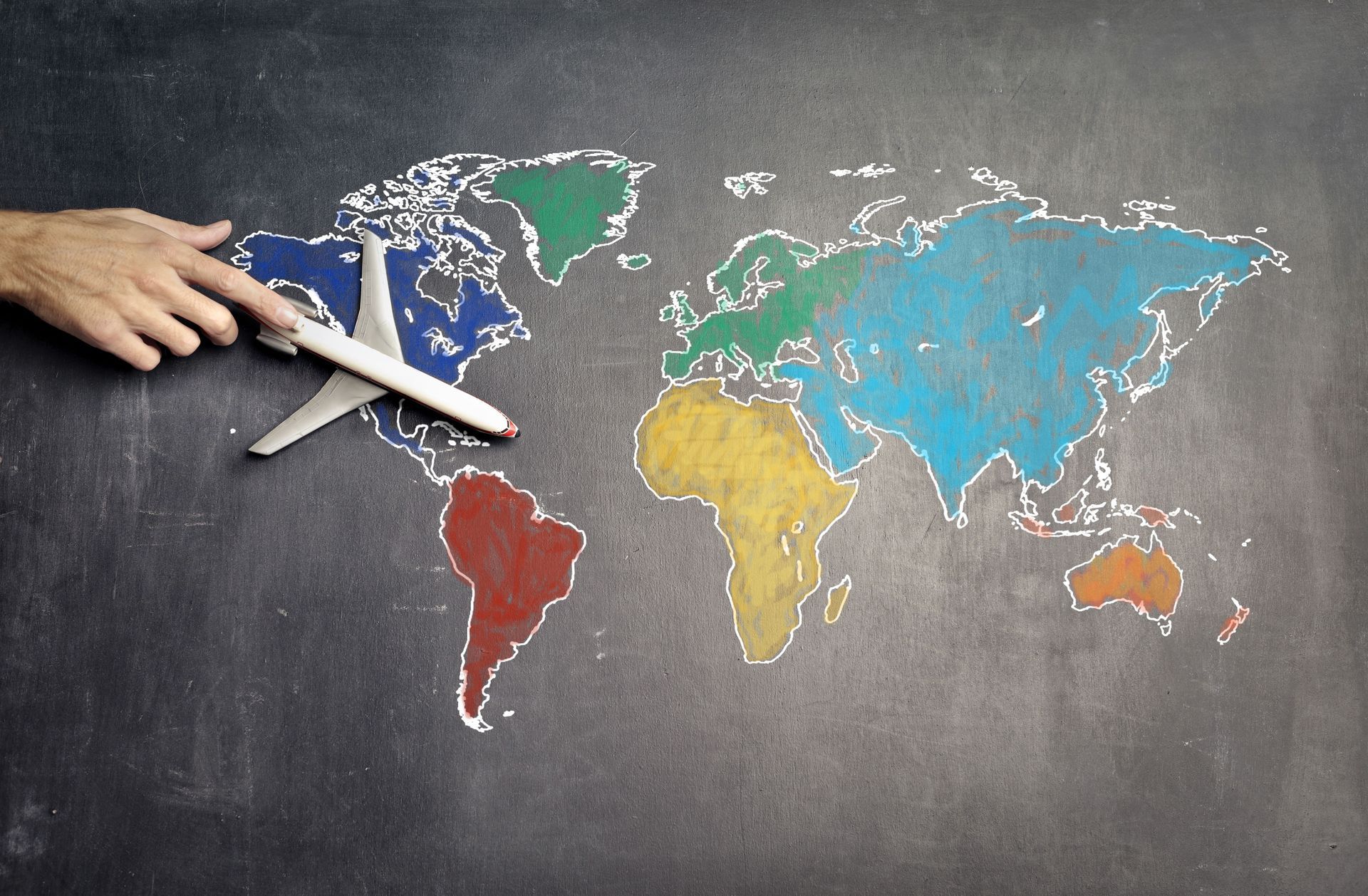 Hand placing a toy airplane over a colorful world map on a chalkboard background