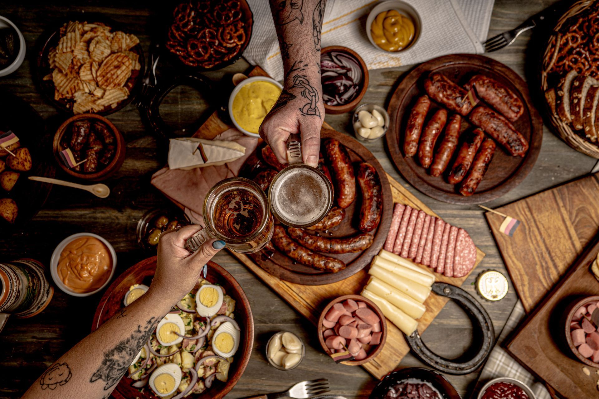Overhead view of a rustic table with hands serving sausages, pretzels, and drinks at a shared meal