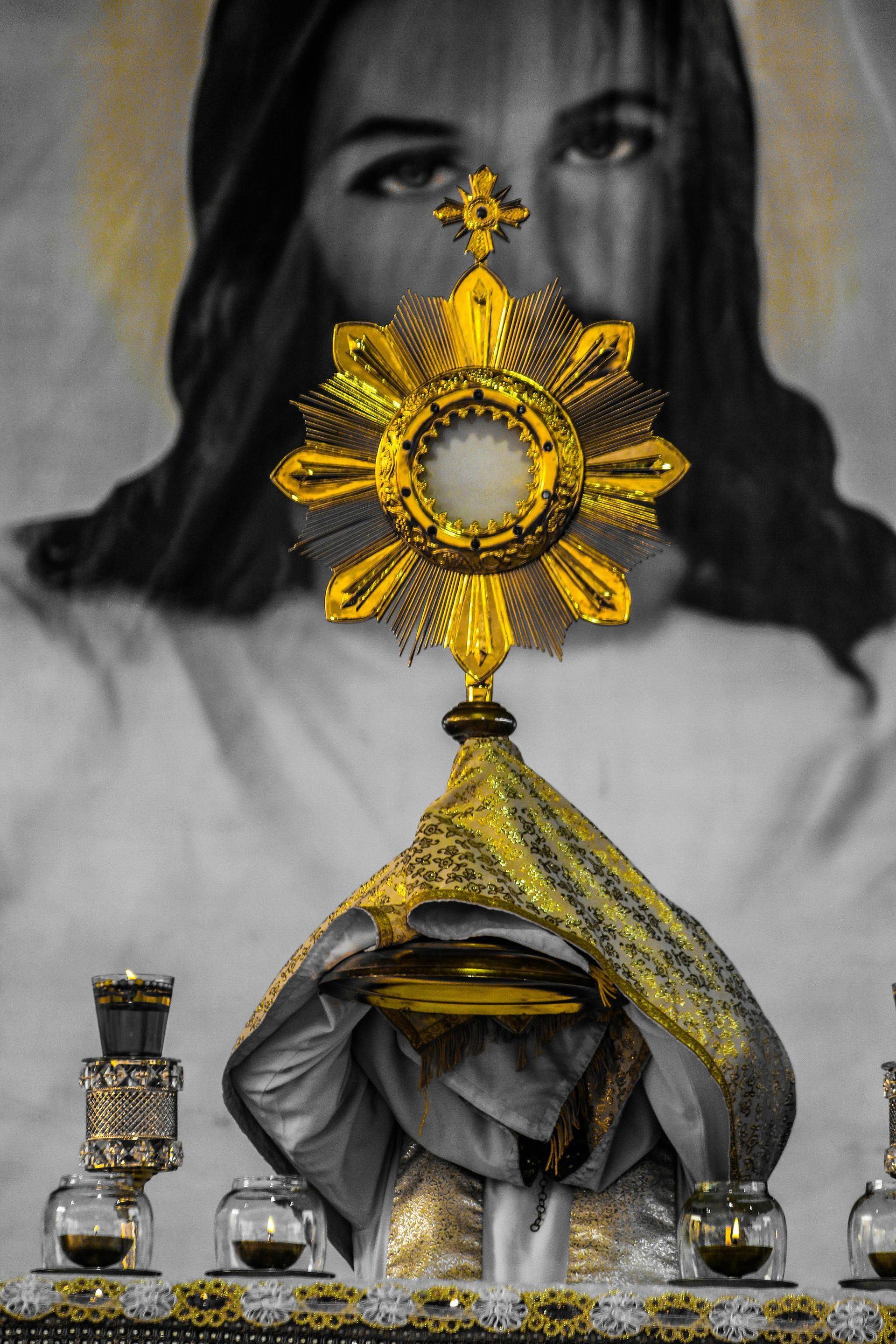 Gold monstrance over a draped statue, with a grayscale portrait in the background.