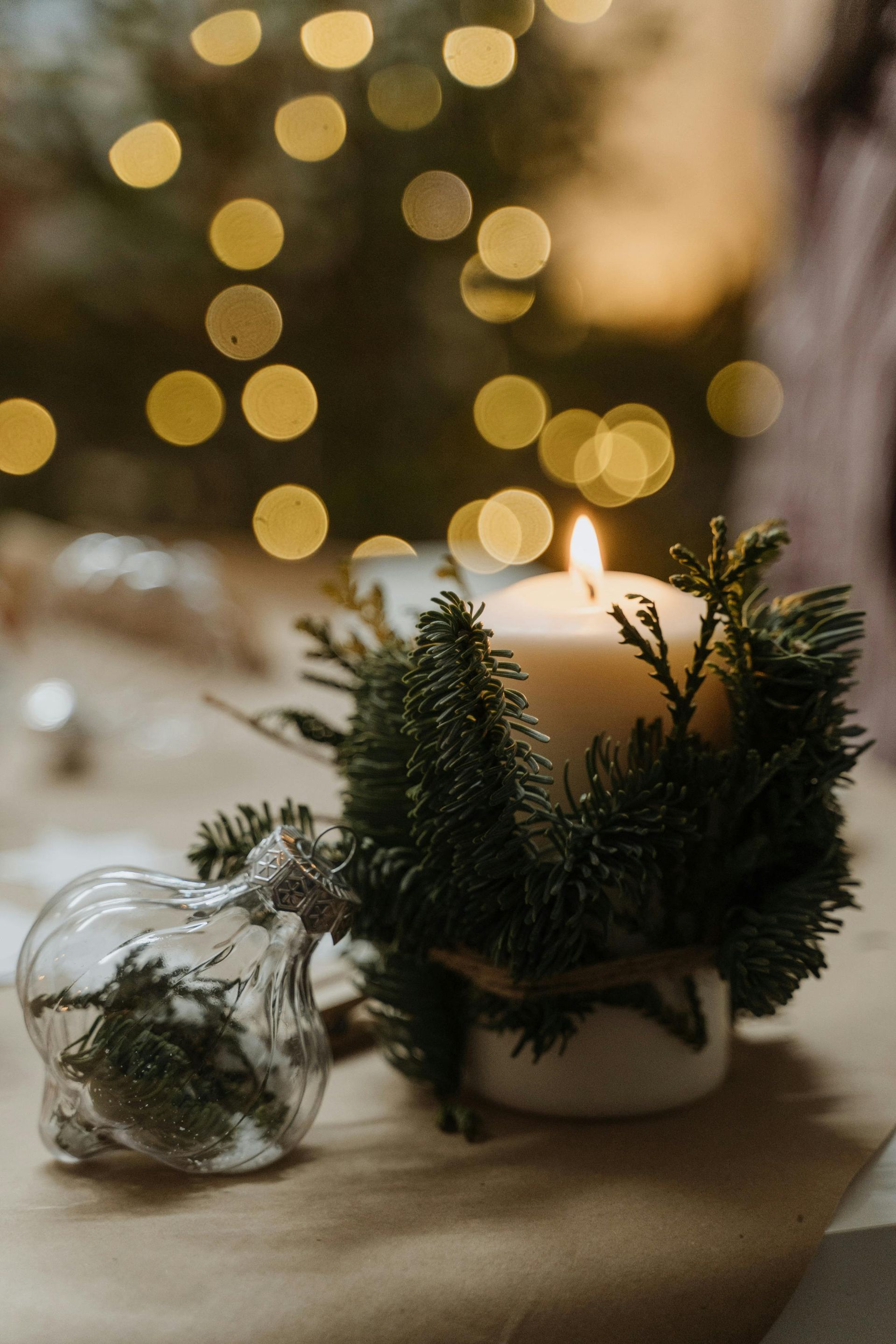 Lit candle in a wreath of greenery; glass ornament beside it. Bokeh lights in the background.
