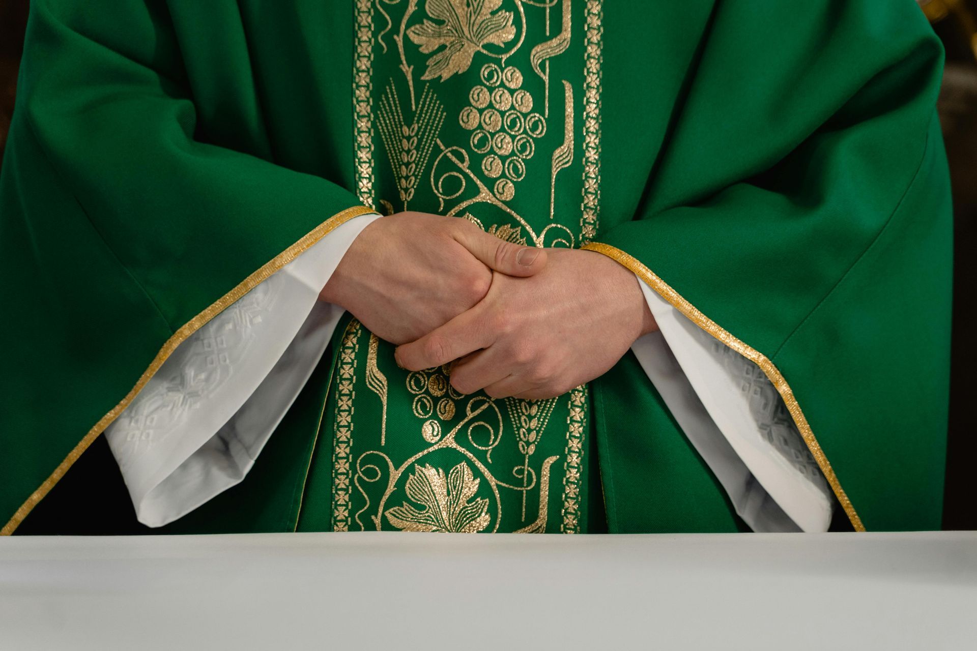 Priest in green liturgical vestments with gold embroidery, hands clasped at a church altar