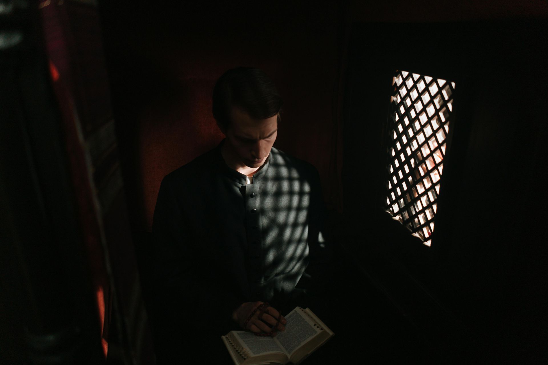 Man in dark confessional reading a book, sunlight through a window.