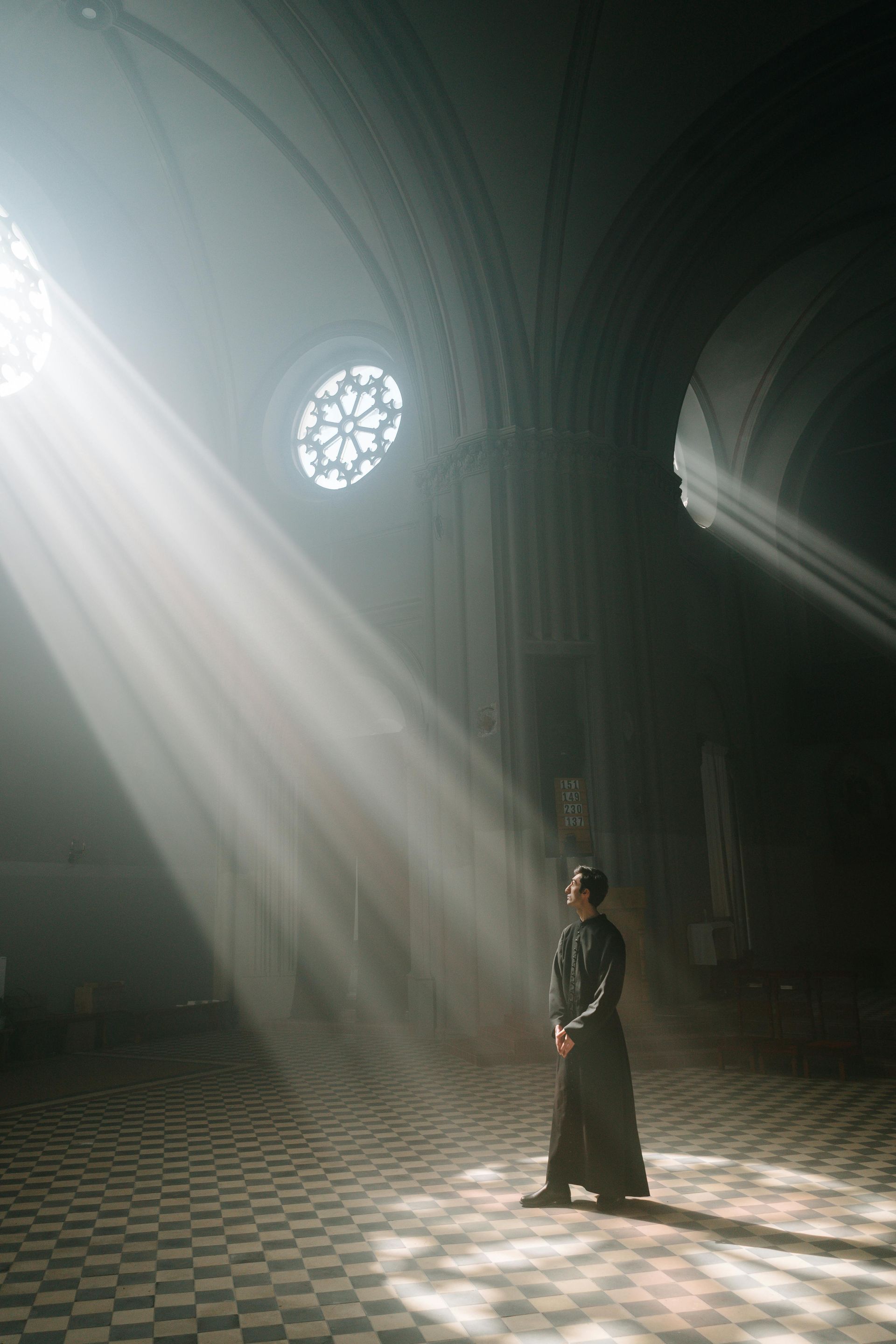 A lone robed figure stands in a dim cathedral, with sunlight streaming through a circular stained-glass window.