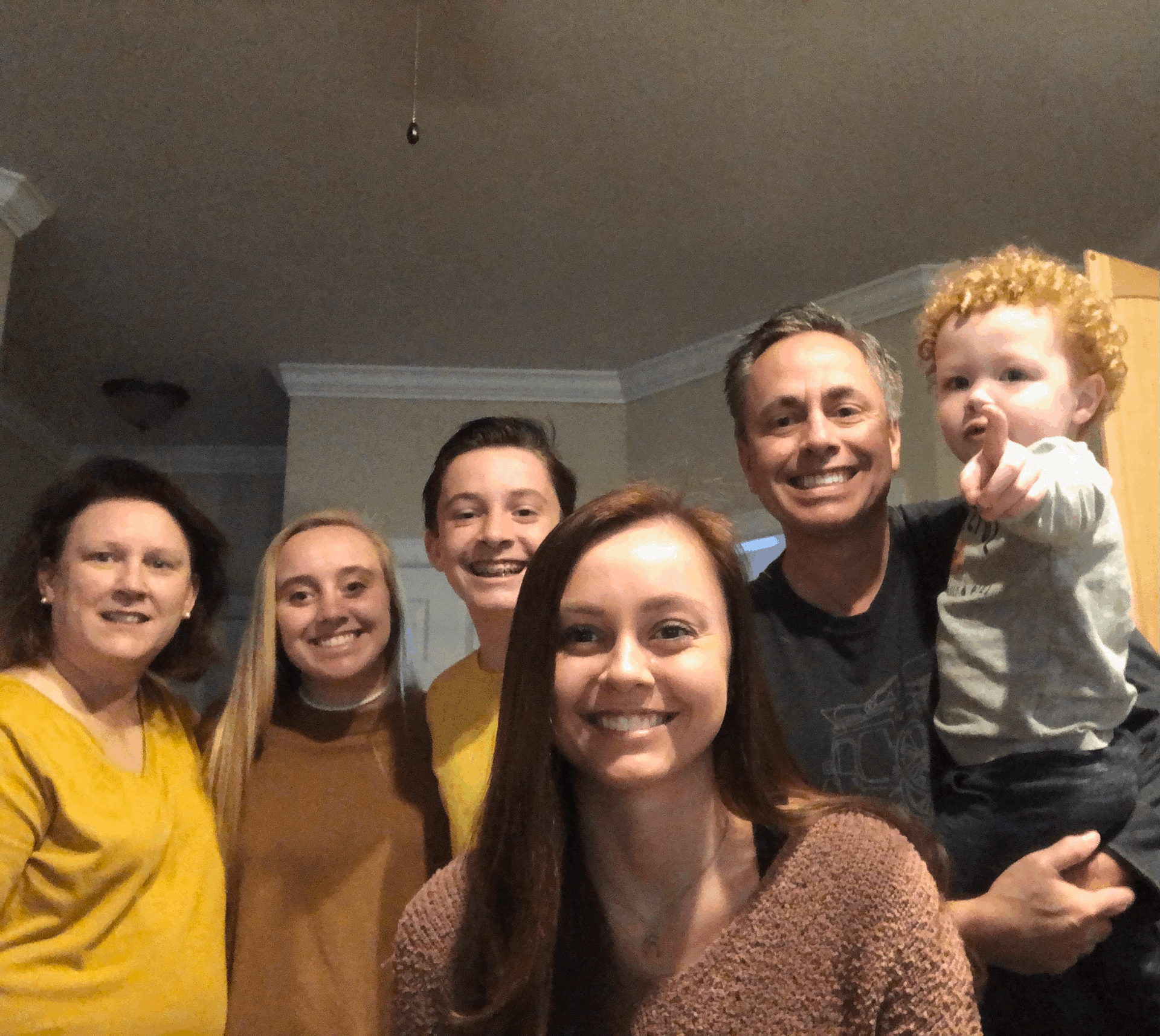 Family smiling indoors, some with arms around each other. The youngest child points towards the camera.