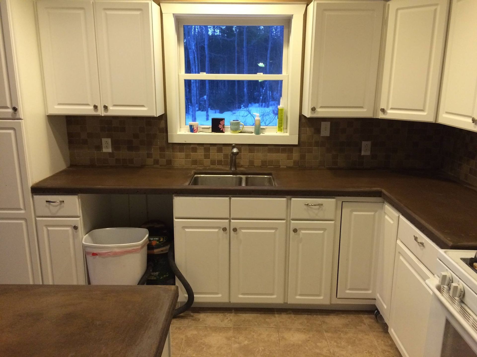 White kitchen with dark countertops, sink under window, white cabinets, and a stove on the right.