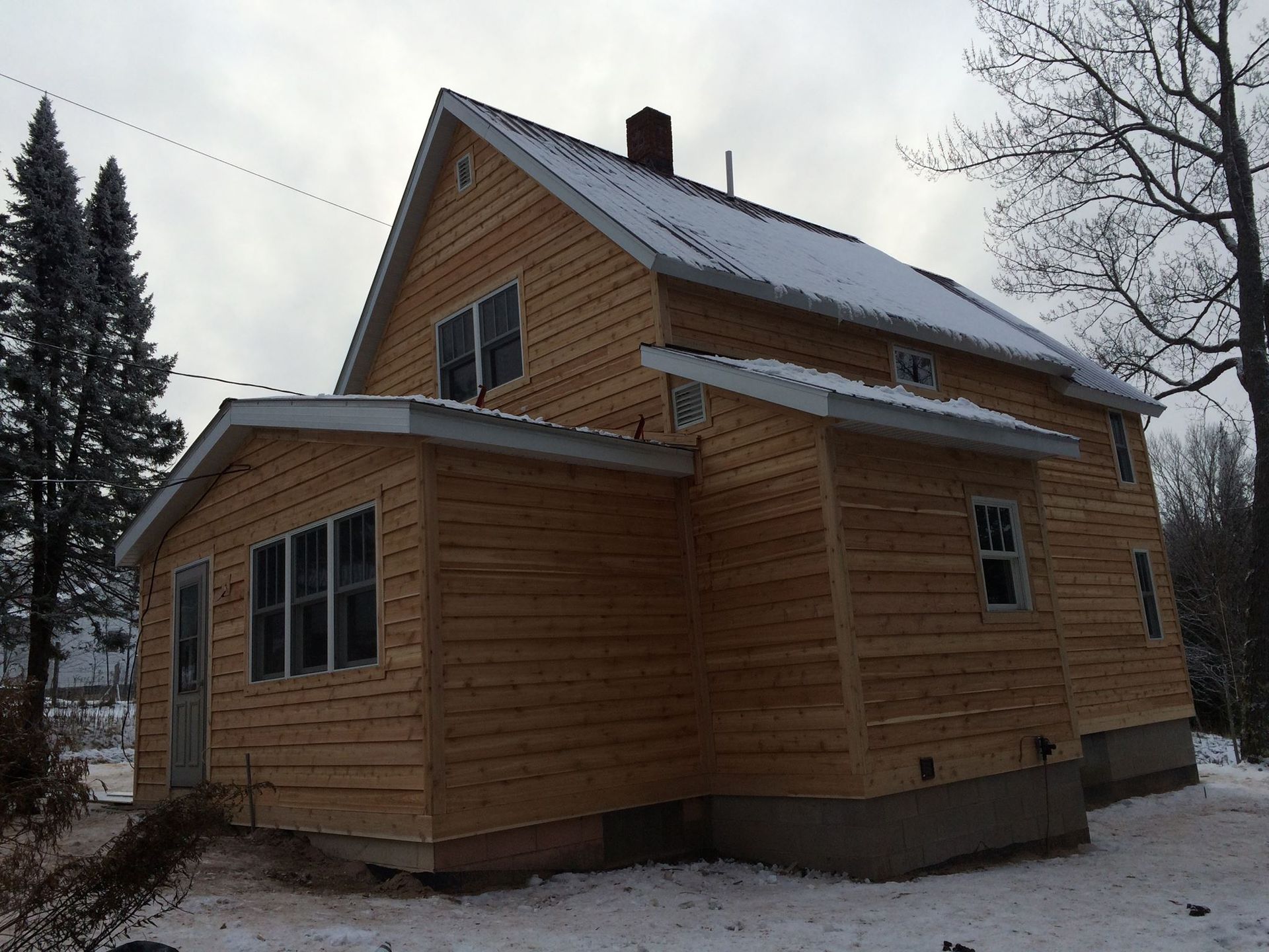 Wooden house with a steep roof in a snowy yard, viewed from the side.