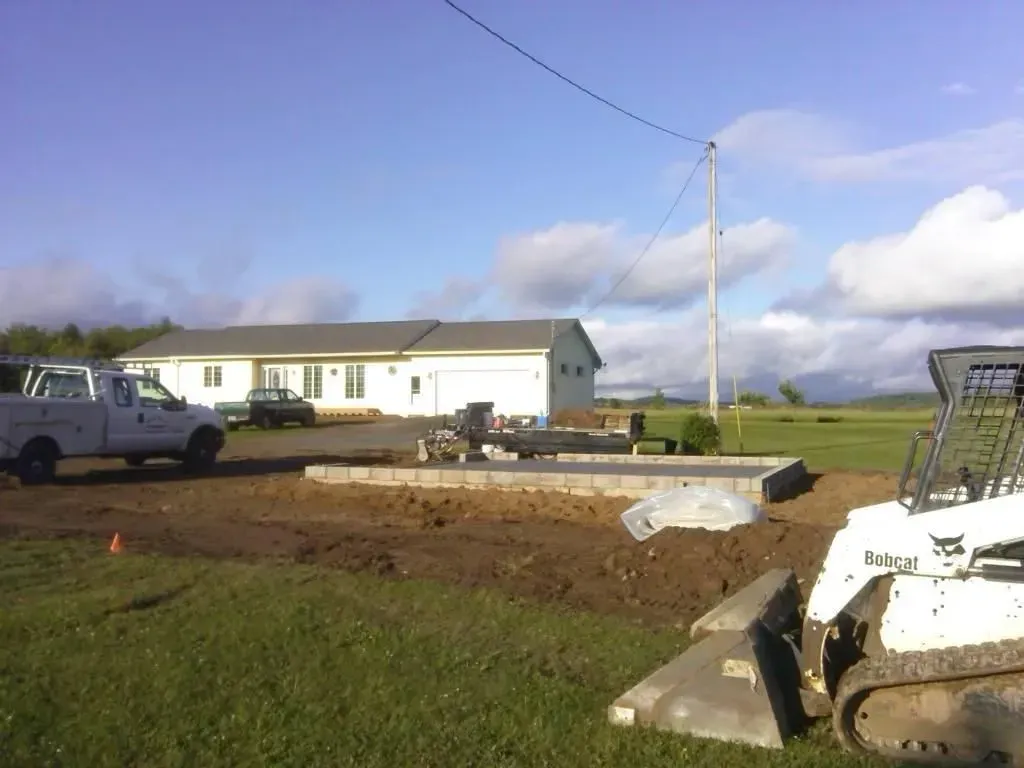 Construction site with a white truck, dirt lot, equipment, and a house under a blue sky