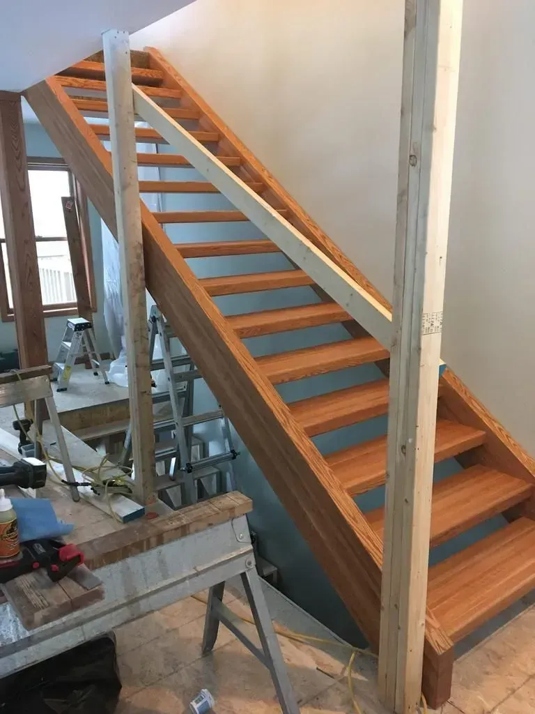 Wooden staircase under construction beside a wall, with support posts and tools in a workshop-like space