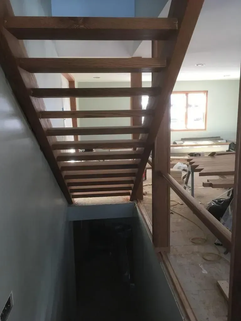 Wooden staircase under construction in a dusty interior room, with a window and tools in the background