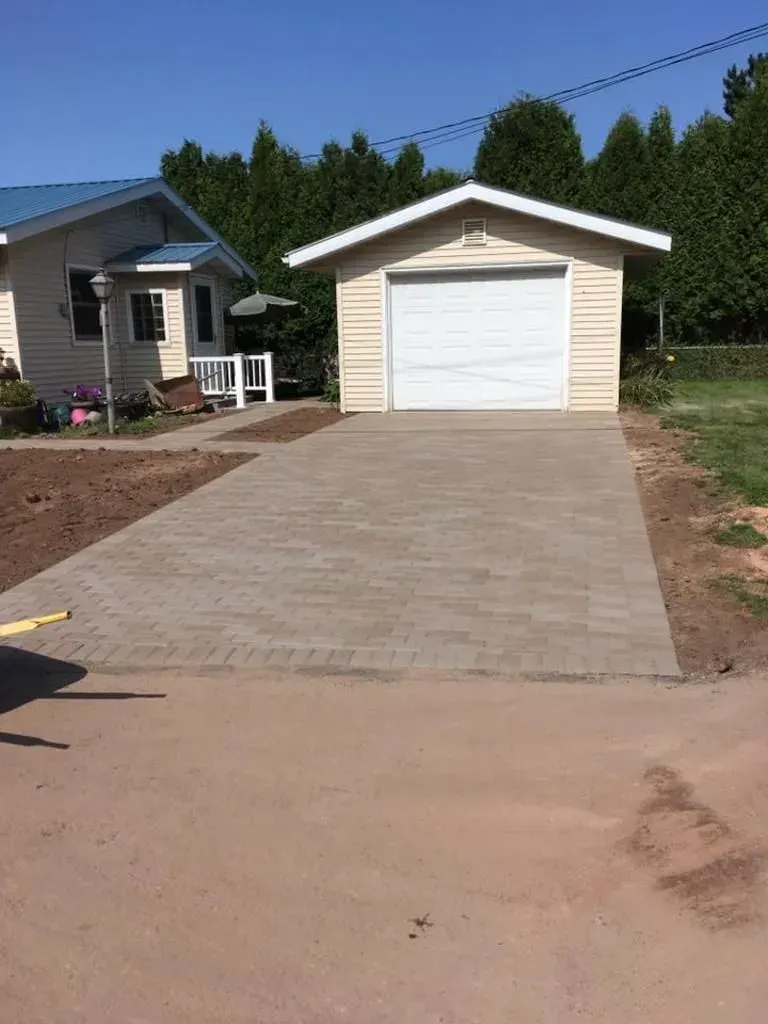 Concrete driveway leading to a white garage beside a small house, with trees in the background.
