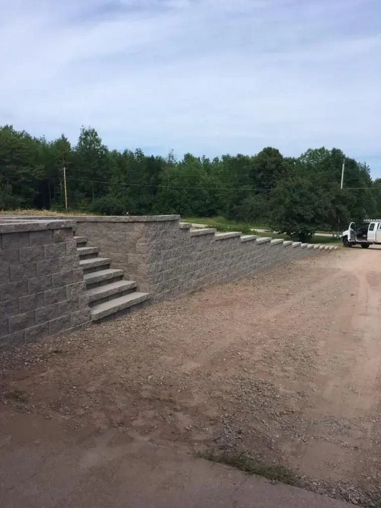 Stone retaining wall with steps beside a dirt lot and trees under a cloudy sky