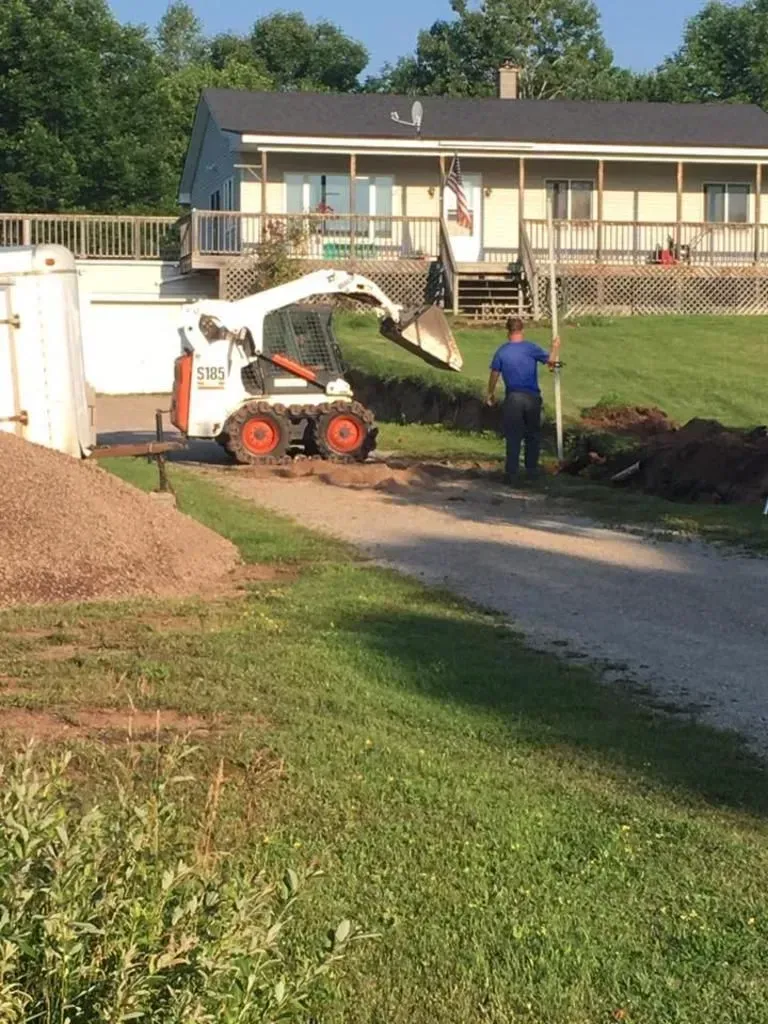 Mini skid steer loading dirt on a gravel driveway beside a house, with a worker nearby.