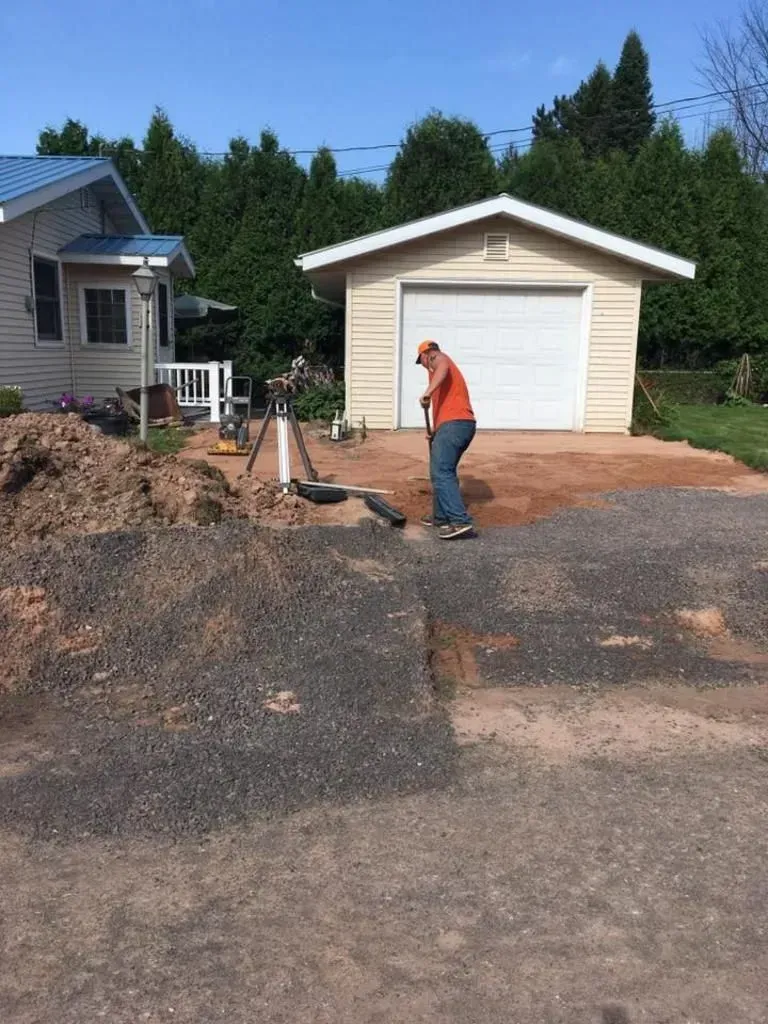 Person shoveling gravel in a driveway beside a house and garage on a sunny day