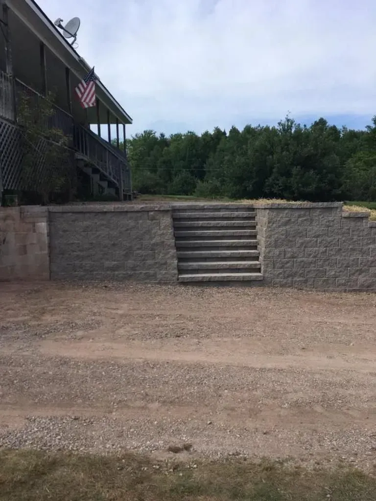 Stone steps lead to a raised patio beside a building with an American flag, overlooking gravel and trees.