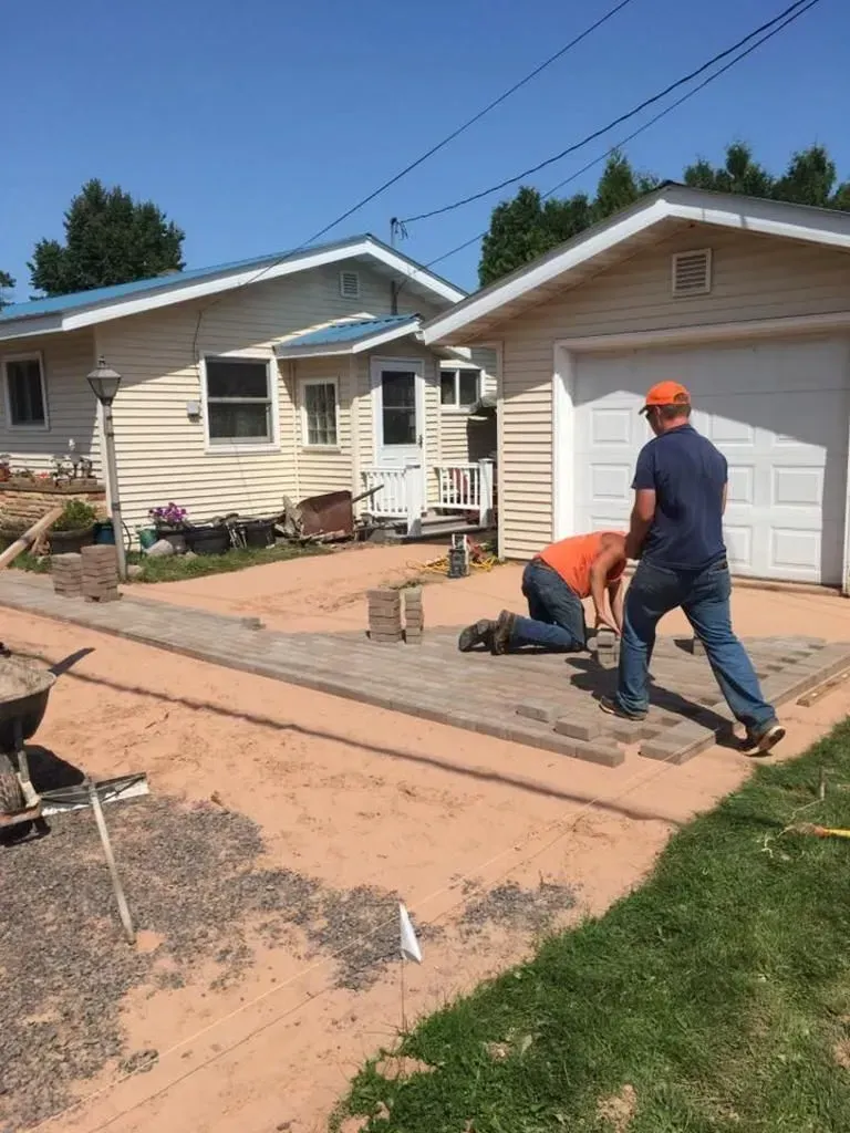 Workers repairing a cracked driveway beside a beige house under a blue sky