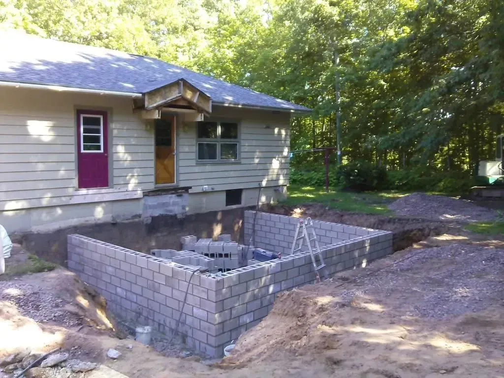 Small gray brick patio or fire pit foundation beside a house in a wooded yard.