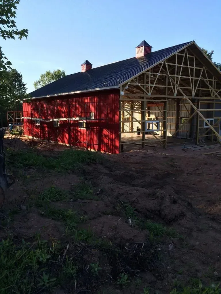 Red barn under construction with exposed wooden framing and a dirt foreground, set beside trees under a blue sky
