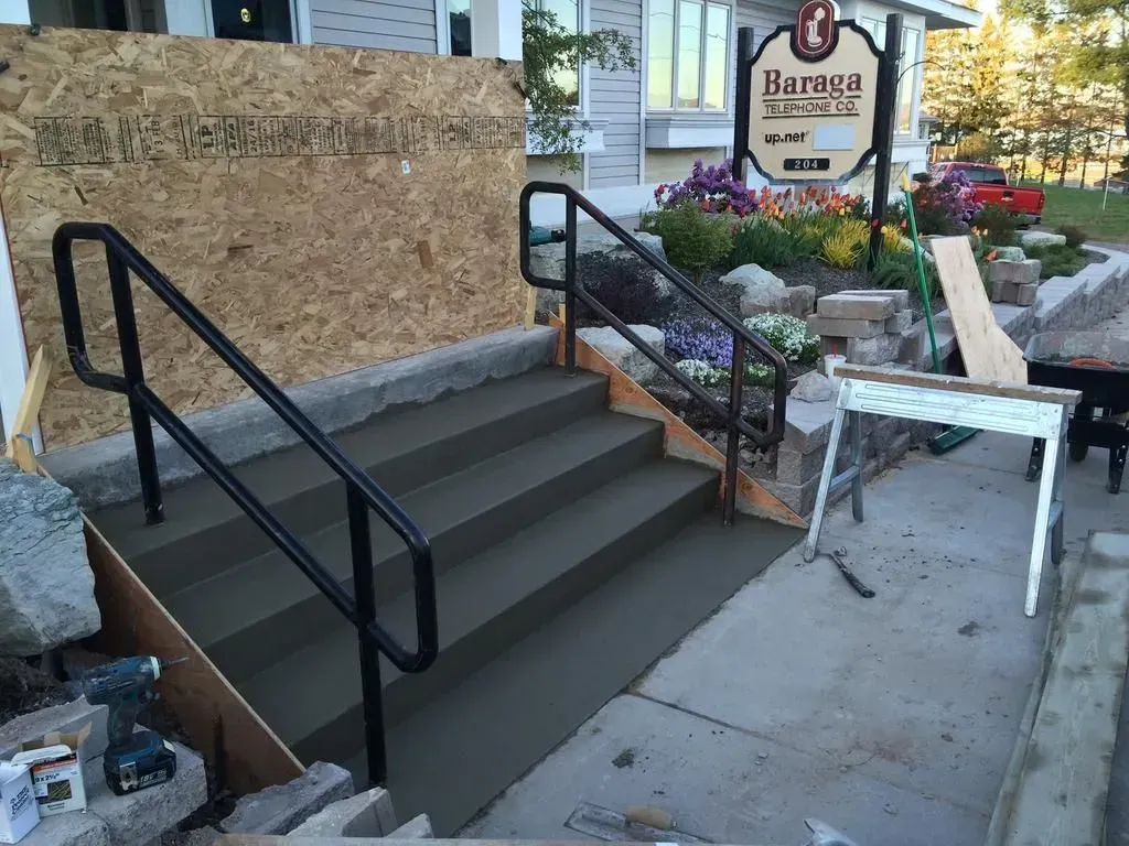 Stone steps with black handrails leading to a storefront entrance beside flowers and a sign.