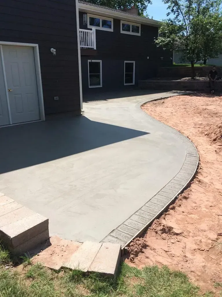 Curved concrete patio beside a dark house, with dirt and a small step at the edge.