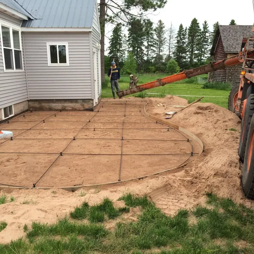 Worker laying concrete patio forms beside a gray house, with sand and rebar grid in the yard.
