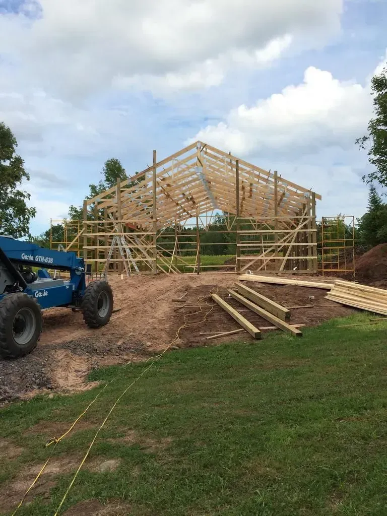 Framed wooden barn under construction with a blue tractor, lumber piles, and scaffolding on a grassy site