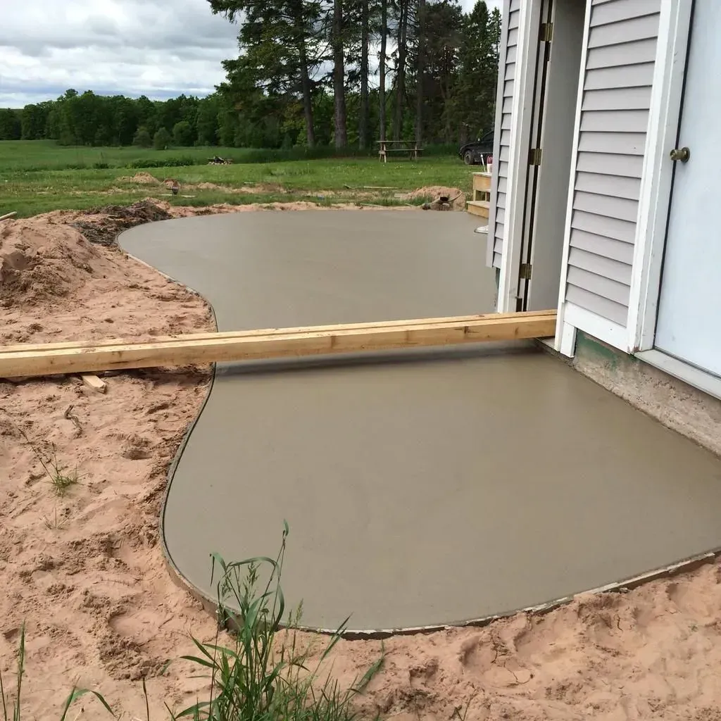 Freshly poured concrete patio beside a house, bordered by dirt and a wooden form board.