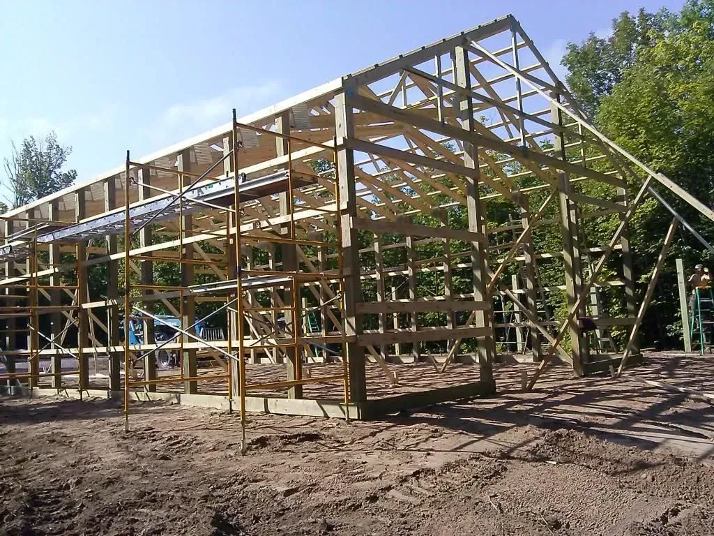 Wooden frame of a house under construction on a dirt lot, with diagonal roof trusses and clear sky.