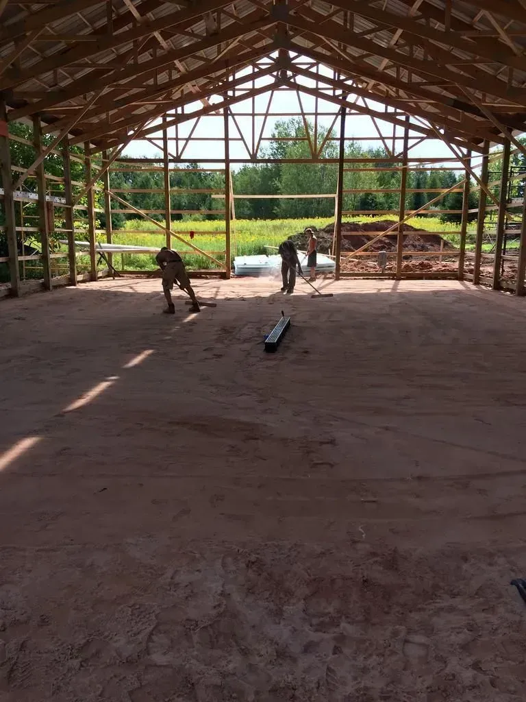 Two workers in a wooden-framed open pavilion, smoothing a dirt floor with tools.