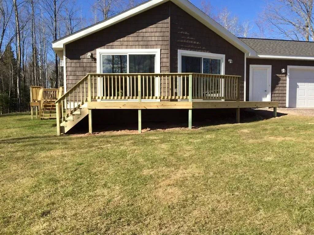 Brown house with a raised wooden deck and stairs, set on a grassy yard under a clear sky