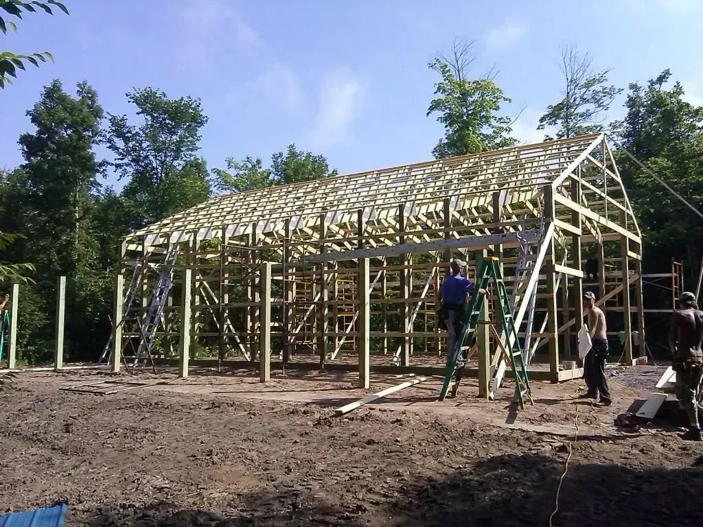 Wooden framework of a building under construction in a wooded outdoor area, with two workers nearby.