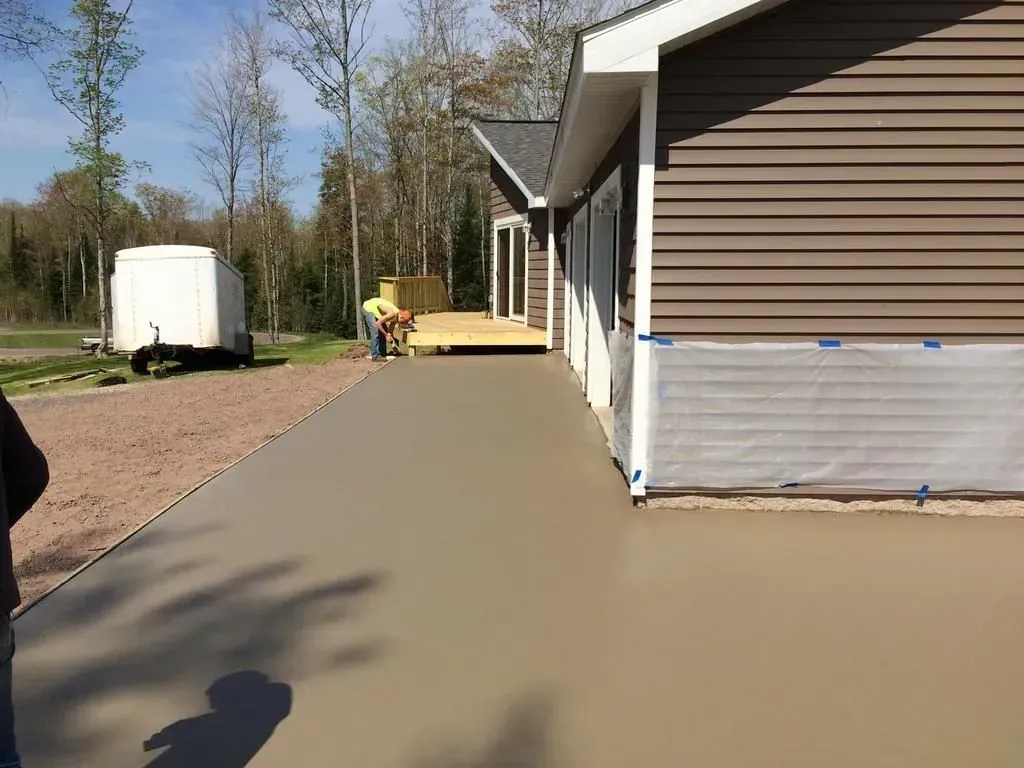 Fresh concrete walkway beside a house under construction, with gravel and a white trailer nearby.