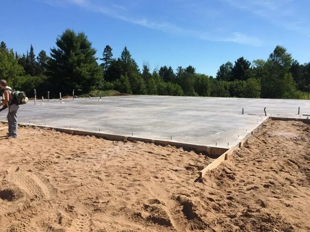 Concrete foundation slab in a sandy lot with trees under a clear blue sky