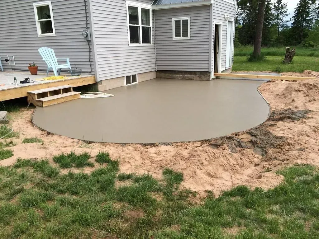 Fresh concrete patio beside a gray house, next to a small wooden deck and muddy yard.