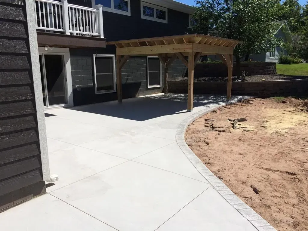 Backyard patio with curved concrete path, wooden pergola, and house exterior beside a dirt yard