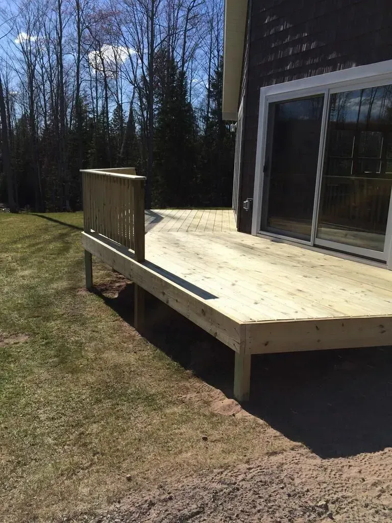 Wooden deck beside a house with sliding glass door, overlooking a grassy yard and trees.
