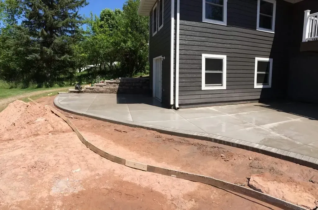 Curved concrete walkway beside a dark house, with exposed dirt and a hose along the edge.