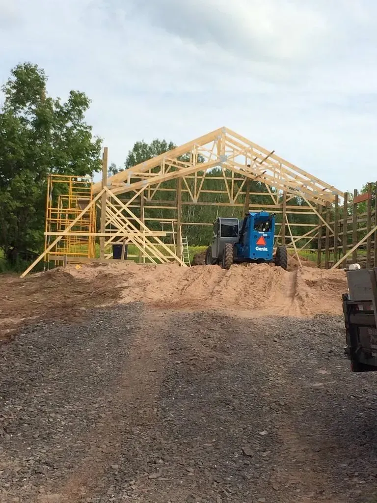 Wooden house frame under construction on a gravel lot, with a blue tractor and trees nearby